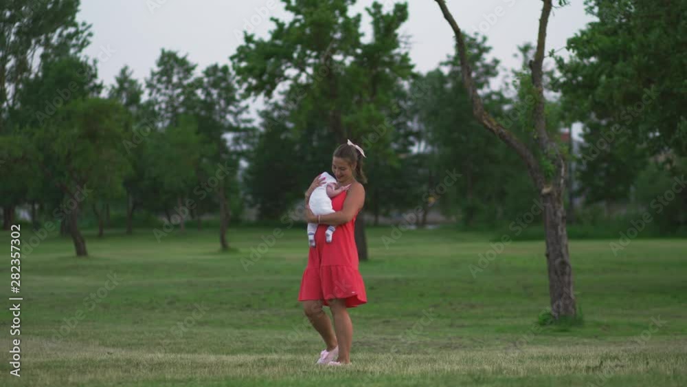 Young mother holding and playing with her baby boy child in city park standing wearing bright red dress - Son wears white cap - Family values warm color summer scene handheld Young mother holding and playing with her baby boy child in city park standing wearing bright red dress - Son wears white cap - Family values warm color summer scene handheld