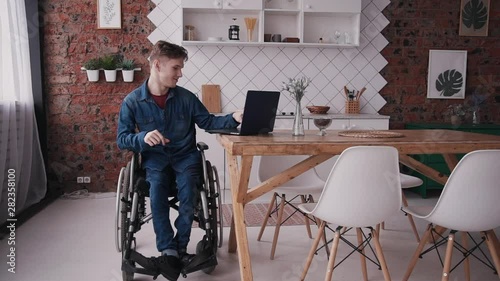 Adult disabled man using his modern computer and spending weekend at cozy bright room in house with interior. Male sitting in special comfort wheelchair making calm glad and smiling face photo