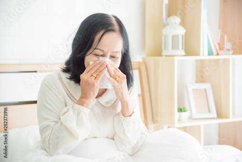 Unwell asian senior woman blowing nose in bed,Senior woman covering nose with tissue paper, sneezing in bed.
