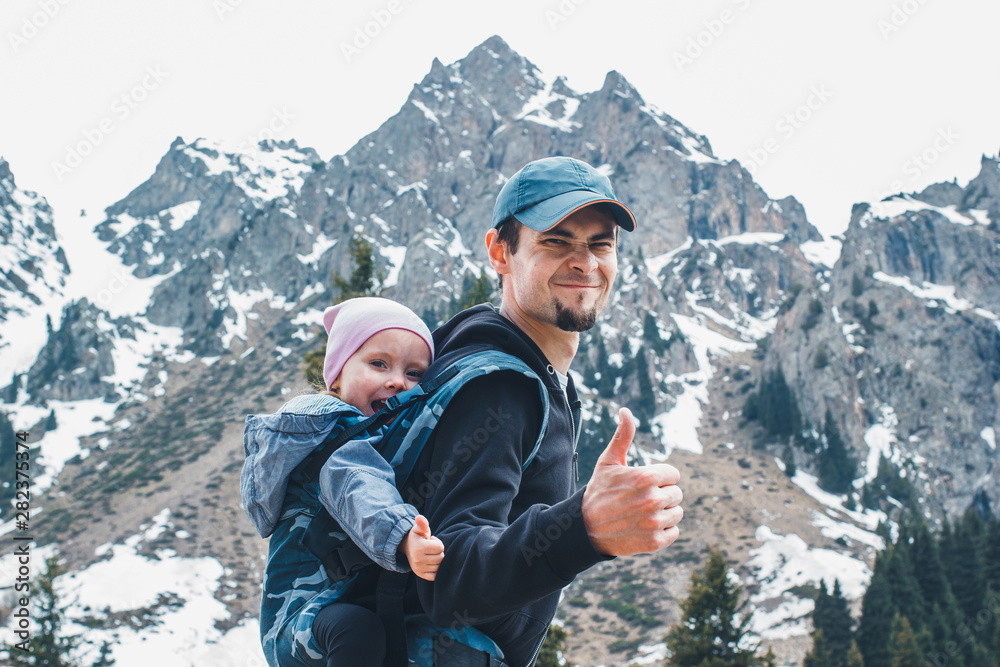 Attractive young father with her baby daughter in ergonomic baby carrier on back outside in mountain nature, show thumbs up.