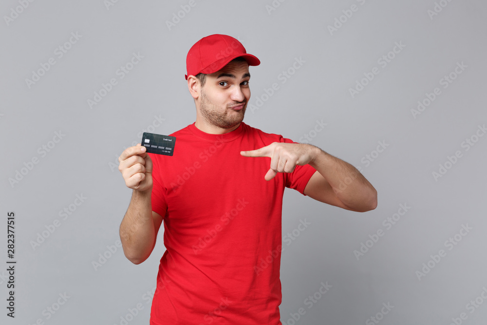 Delivery man in red uniform hold credit card isolated on grey wall background, studio portrait. Professional male employee in cap t-shirt print working as courier. Service concept. Mock up copy space.