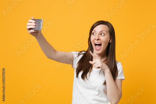 Portrait of excited young woman in white casual clothes keeping mouth open, doing selfie shot on mobile phone isolated on bright yellow orange background. People lifestyle concept. Mock up copy space.