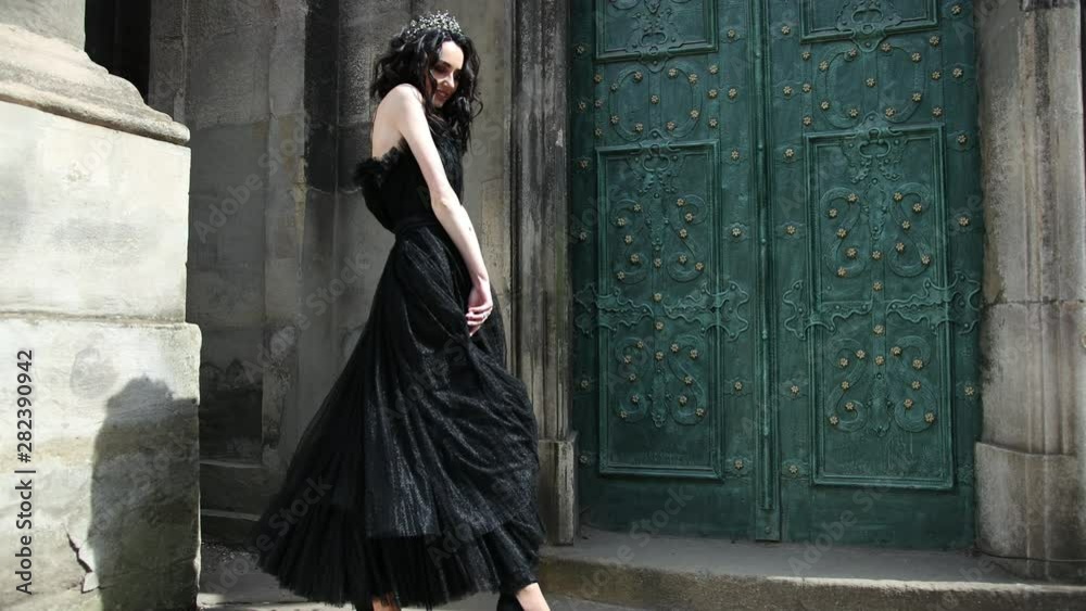 thin beautiful girl, brunette, with light skin, in a long black dress, with bright makeup, crown on his head, posing, smiling, on the street near the old building, with high green doors, slow shooting