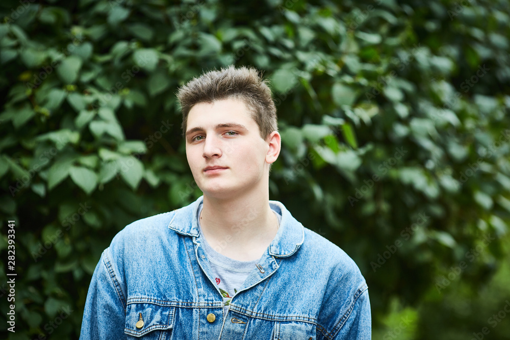 Closeup of a young handsome serious Caucasian guy in a denim jacket on a background of foliage Closeup of a young handsome serious Caucasian guy in a denim jacket on a background of foliage