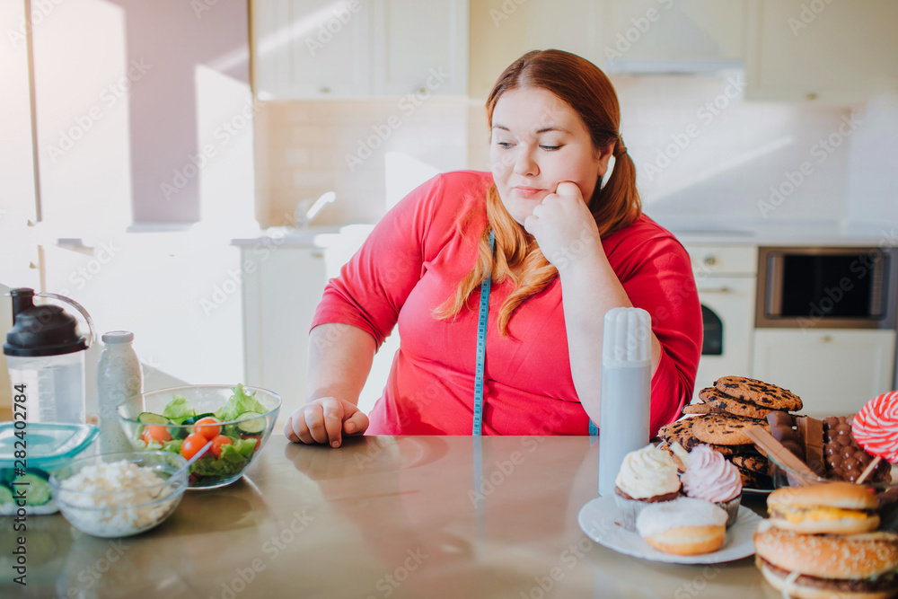 Fat young woman in kitchen sitting and eating food. She look at healthy meal on left side. Soft measure tape around neck. Sweet junk food on right side. Daylight in kitchen. Fat young woman in kitchen sitting and eating food. She look at healthy meal on left side. Soft measure tape around neck. Sweet junk food on right side. Daylight in kitchen.