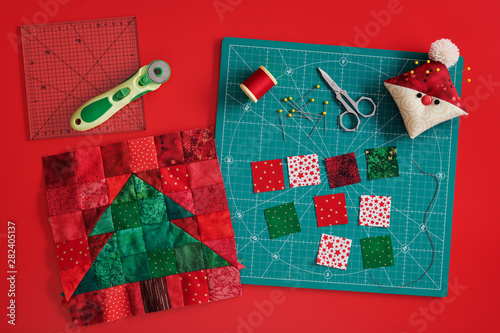 Christmas tree patchwork block, craft mat, bright square pieces of fabric, pincushion like Santa and quilting accessories on red background