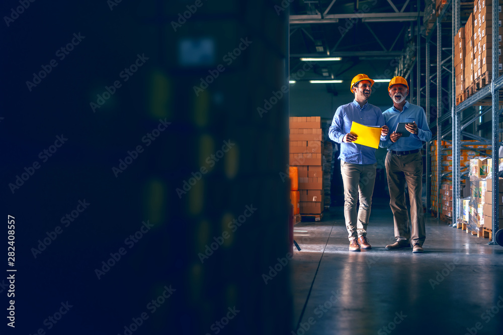 CEO going around warehouse with supervisor and talking analyzing sale statistics. Younger man holding folder with data while older one holding tablet. Both having yellow helmets.