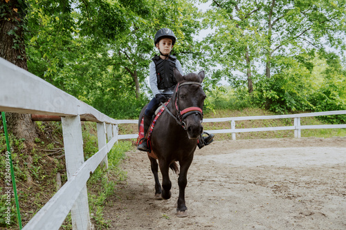 Playful little jockey boy riding adorable pony at sunny day on ranch.