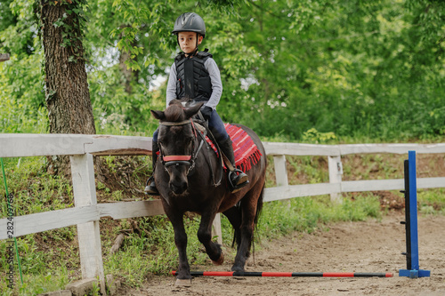 Playful little jockey boy riding adorable pony at sunny day on ranch.