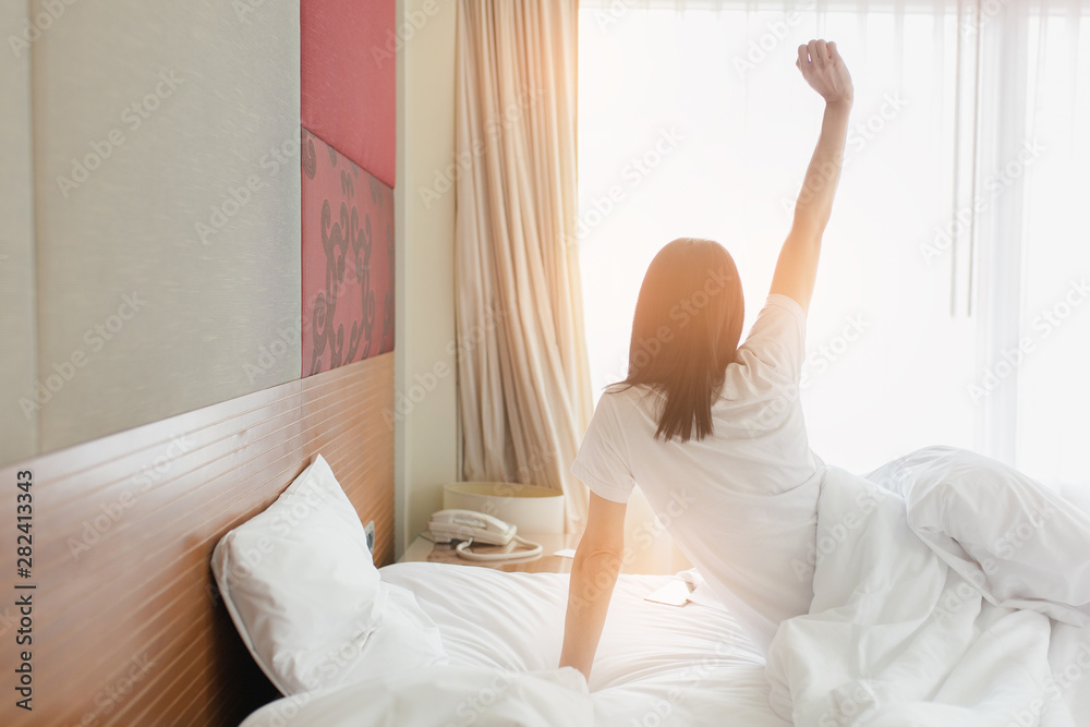 Woman stretching in bed after waking up, back view. Woman sitting near the big white window while stretching on bed after waking up with sunrise at morning, back view. Woman stretching in bed after waking up, back view. Woman sitting near the big white window while stretching on bed after waking up with sunrise at morning, back view.