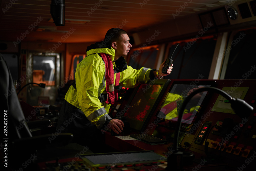 Navigator. pilot, captain as pat of ship crew performing daily duties with VHF radio, binoculars on board of modern ship with high quality navigation equipment on the bridge on sunrise. Navigator. pilot, captain as pat of ship crew performing daily duties with VHF radio, binoculars on board of modern ship with high quality navigation equipment on the bridge on sunrise.