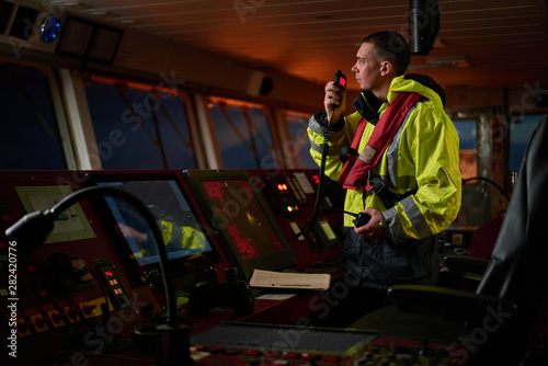 Navigator. pilot, captain as pat of ship crew performing daily duties with VHF radio, binoculars on board of modern ship with high quality navigation equipment on the bridge on sunrise.