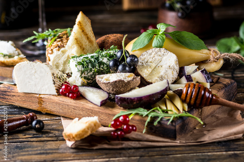 Cheese appetizer selection. Red currant, honey, basil, grapes and nuts on rustic wooden board over wooden concrete backdrop, top view. Belper Knolle, goat cheese, Scamorza. Cheese board, cheese plate