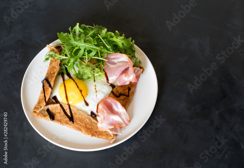 Tableau sur toile Galette bretonne  with fried eggs, arugula and bacon on a black background