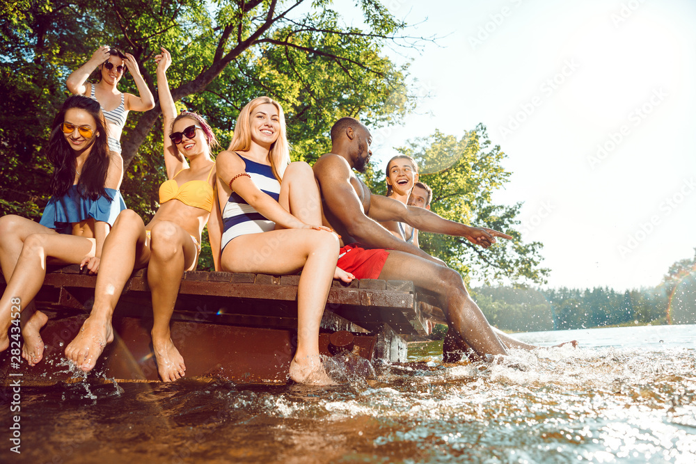 Fototapeta premium Group of happy friends having fun while splashing water and laughting on the pier on river. Joyful women and men in swimsuit at riverside in sunny day. Summertime, friendship, resort, relations