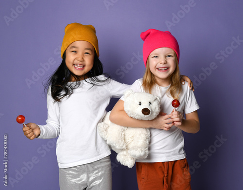 Fototapeta Naklejka Na Ścianę i Meble -  Two laughing kid girls in white t-shirt and colorful hats hold polar bear toy and lollipop candy on purple