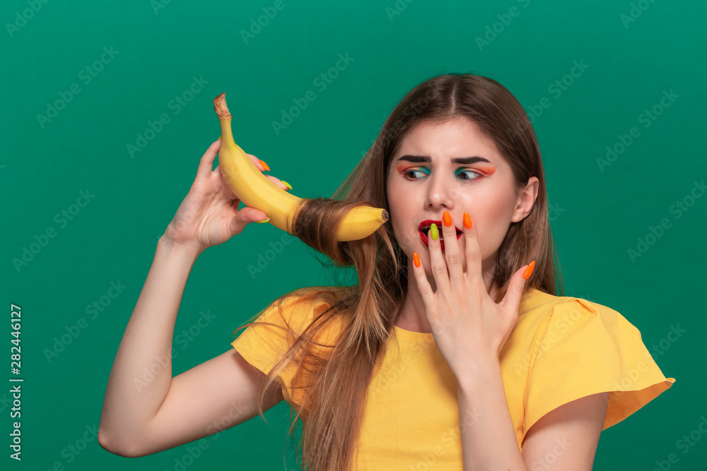 Fototapeta premium Portrait of beautiful young woman with bright color make-up jokingly curling hair with banana.