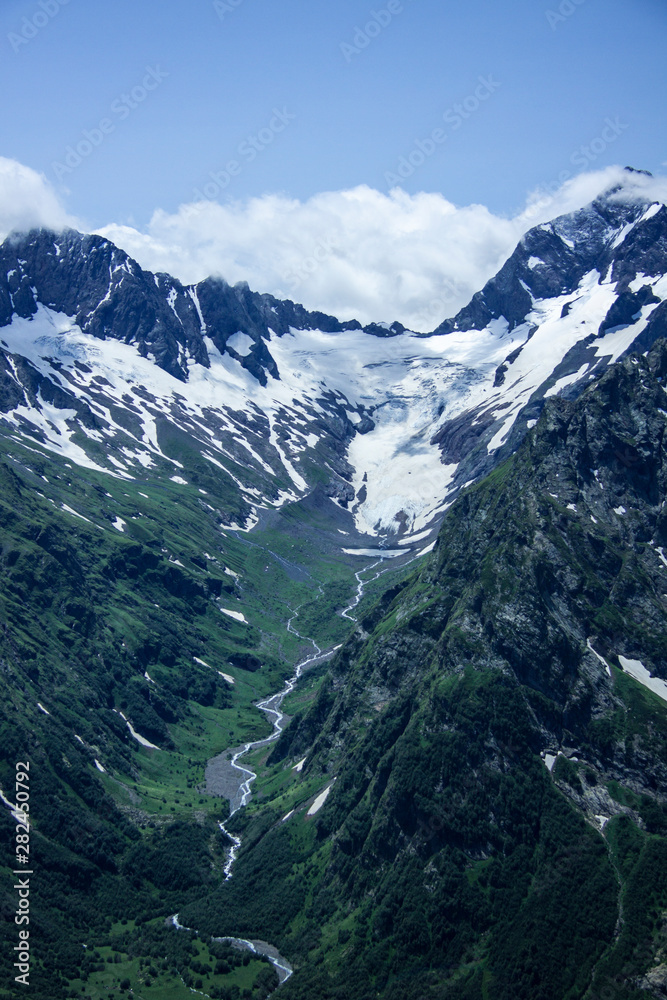 Vertical beautiful panorama of the Caucasus mountains. The top of the mountain range covered with snow. Forest on the slope. Sunny day. Background image for travel and nature.