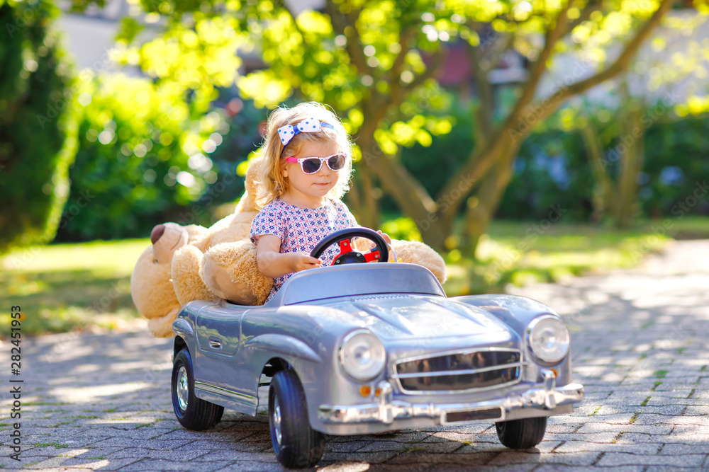 Little adorable toddler girl driving big vintage toy car and having fun with playing with plush toy bear, outdoors. Gorgeous happy healthy child enjoying warm summer day. Smiling stunning kid in gaden