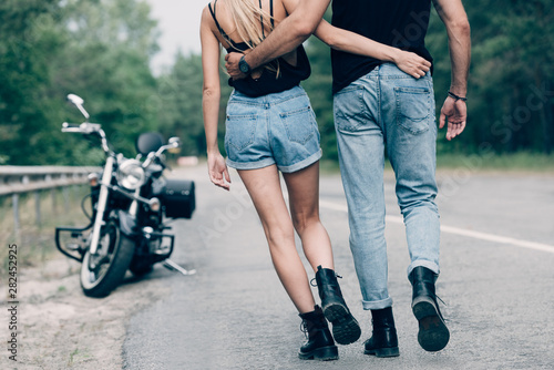 partial view of young couple of bikers walking along road and embracing near black motorcycle