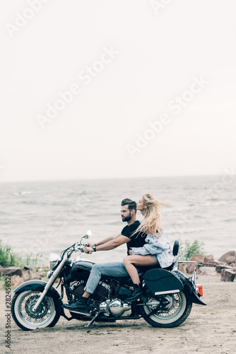 side view of young couple of bikers riding black motorcycle at sandy beach near river