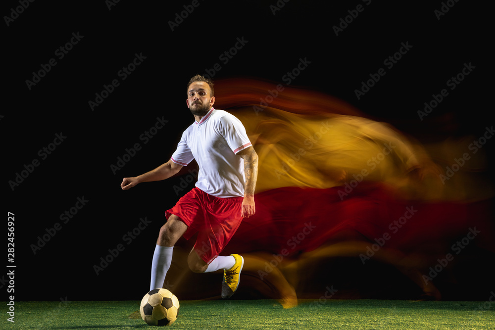 Propose. Young caucasian male football or soccer player in sportwear and boots kicking ball for the goal in mixed light on dark background. Concept of healthy lifestyle, professional sport, hobby.