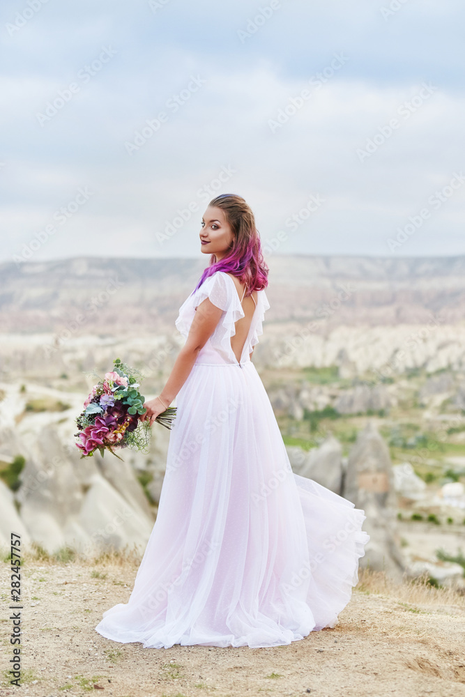 Woman with a beautiful bouquet of flowers in her hands stands on the mountain in the rays of the dawn sunset. Beautiful white long dress on the girl body. Perfect bride with pink hair