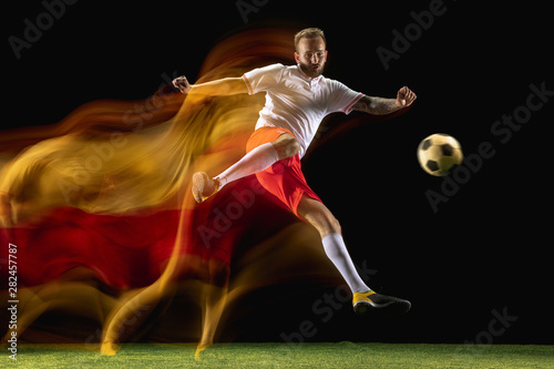 Confident. Young caucasian male football or soccer player in sportwear and boots kicking ball for the goal in mixed light on dark background. Concept of healthy lifestyle, professional sport, hobby.