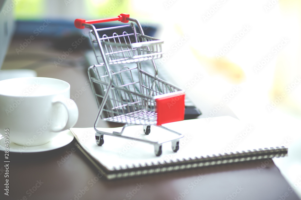 Online shopping concept.Shopping cart on desk with notebook and white cup of coffee