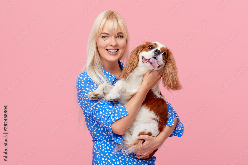 Portrait of smiling young blond woman in summer hat embracing king Charles spaniel dog. owner and pet relations concept. Veterinary health. Isolated front view on pink background. Portrait of smiling young blond woman in summer hat embracing king Charles spaniel dog. owner and pet relations concept. Veterinary health. Isolated front view on pink background.