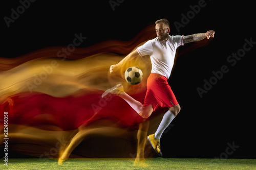 Young caucasian male football or soccer player in sportwear and boots kicking ball for the goal in mixed light on dark background. Concept of healthy lifestyle  professional sport  hobby.