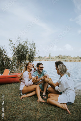 Group of friends with cider bottles sitting by the boat near the beautiful lake and having fun