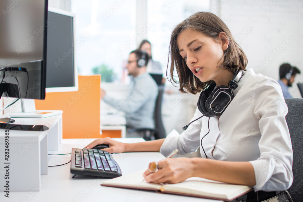 Young business woman working in front of computer, writing notes to notebook while sitting at her workplace in office Young business woman working in front of computer, writing notes to notebook while sitting at her workplace in office
