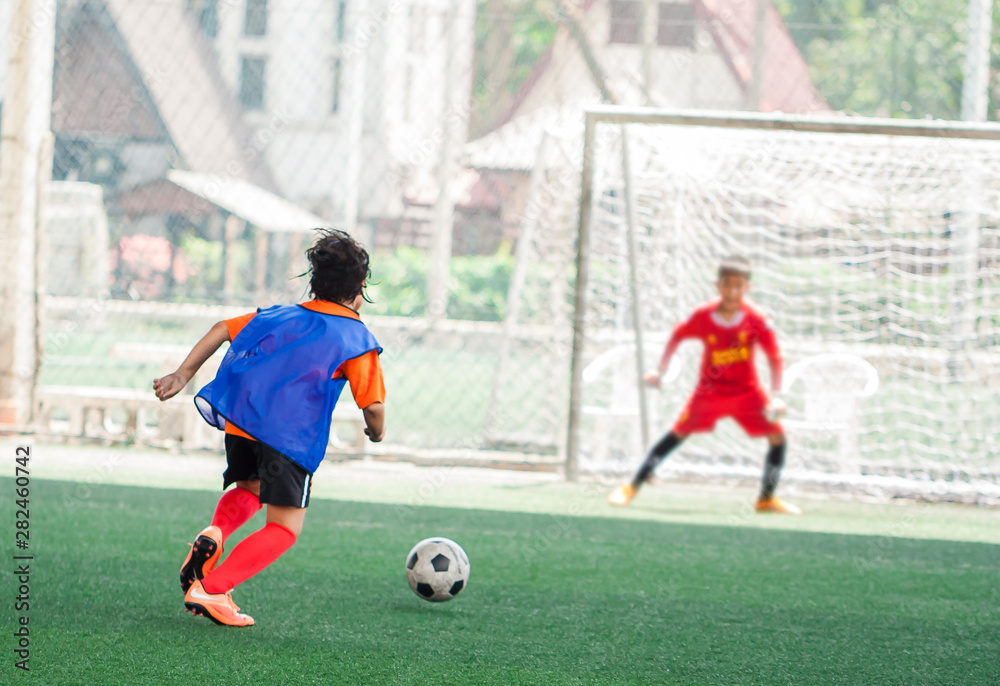 Obraz premium Kid soccer player run to shoot ball to goal with blurry goalkeeper background on green artificial turf, Football or soccer academy