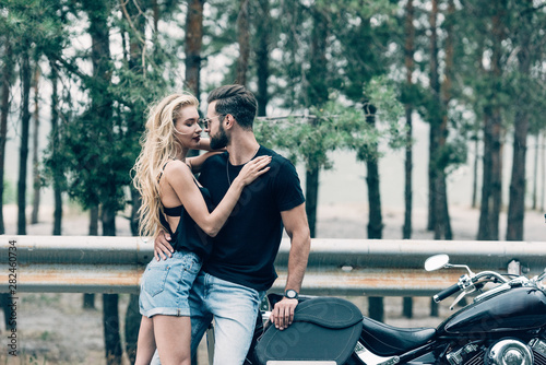 young couple of bikers tenderly embracing near black motorcycle on road near green forest