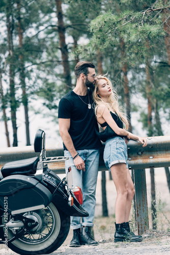 young couple of bikers embracing near black motorcycle on road near green forest