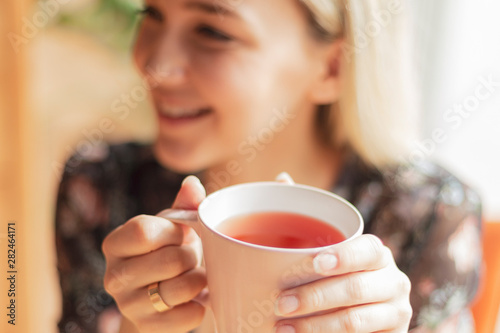 Close up photo of pretty woman holding mug of delicious hot tea, drinking, smiling, in cozy morning cafe, selective focus, noise effect