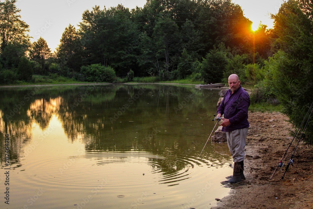 Peaceful Scene of Middle Aged Man Fishing on a Country Pond at Sunset with Golden filtered light reflected on pond