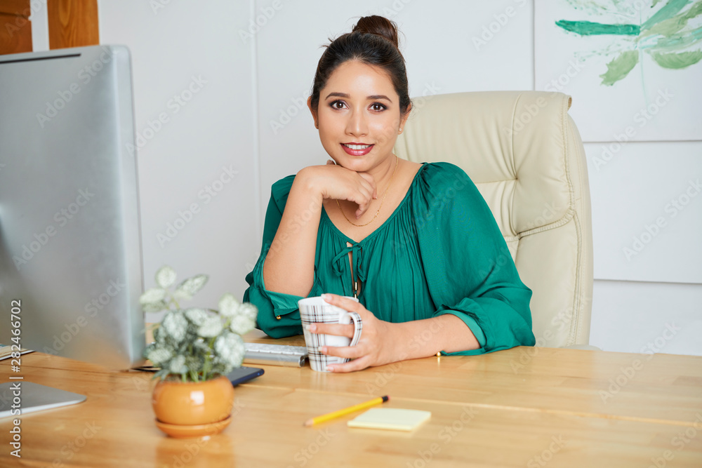 Portrait of Indian beautiful businesswoman sitting at the table in front of computer drinking coffee and smiling at camera at office Portrait of Indian beautiful businesswoman sitting at the table in front of computer drinking coffee and smiling at camera at office