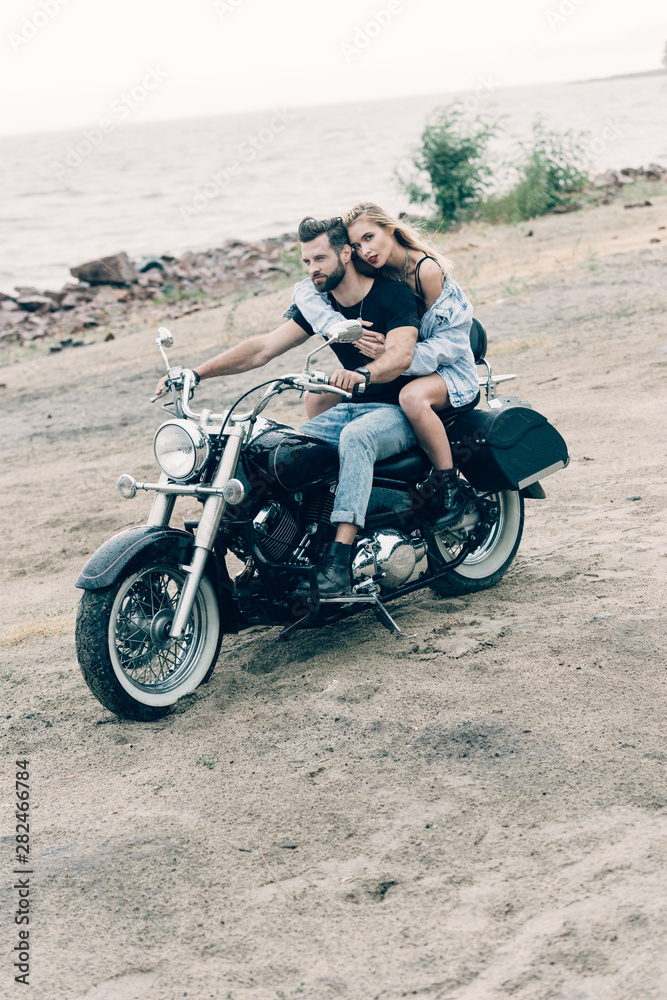 young couple of bikers riding black motorcycle at sandy beach