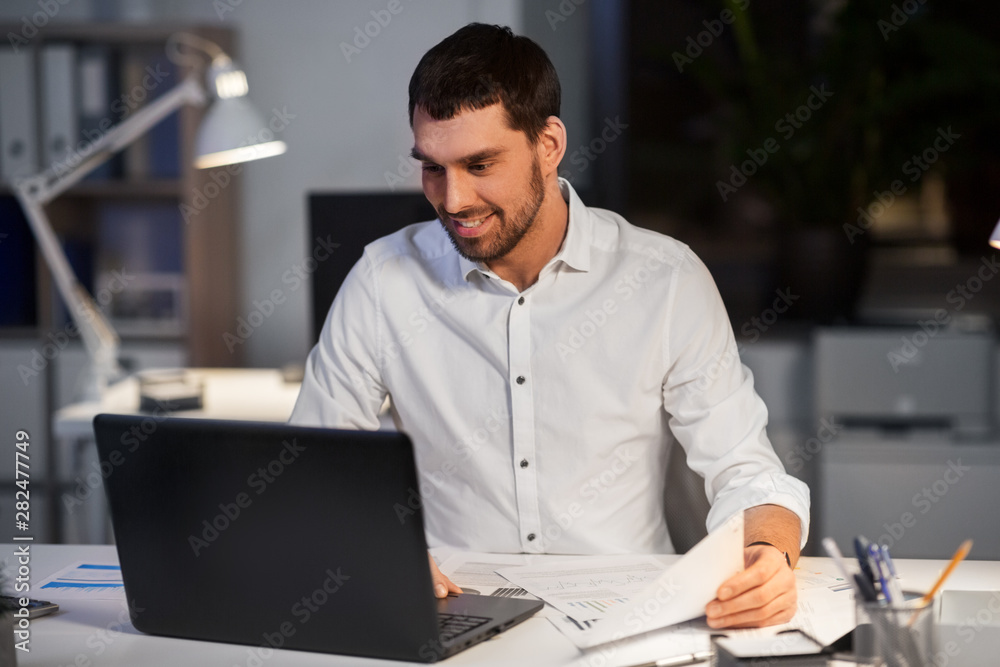 Obraz premium business, deadline and people concept - businessman with laptop computer and papers working at night office