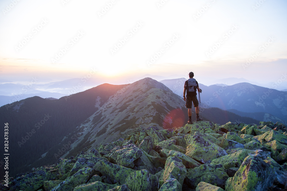 caucasian man hiker with backpack and trekking sticks is on the peak of green stones mountain enjoy a panoramic view of mountains in the sunset light