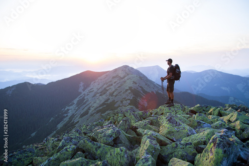 caucasian man hiker with backpack and trekking sticks is on the peak of green stones mountain enjoy a panoramic view of mountains in the sunset light © AlexR