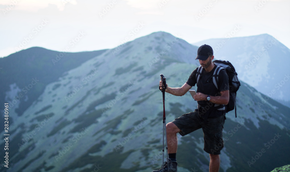 caucasian man hiker with backpack and trekking sticks is on the peak of green stones mountain is searching something in the smartphone