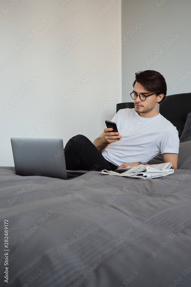 Fototapeta premium Young handsome serious brunette man in white T-shirt and eyeglasses thoughtfully using cellphone lying on big bed with laptop and newspapers at home