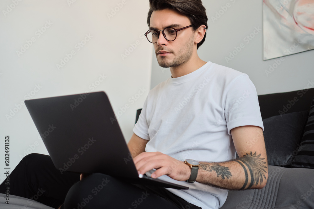 Young attractive brunette man in T-shirt and eyeglasses sitting on bed at modern home and thoughtfully using on laptop