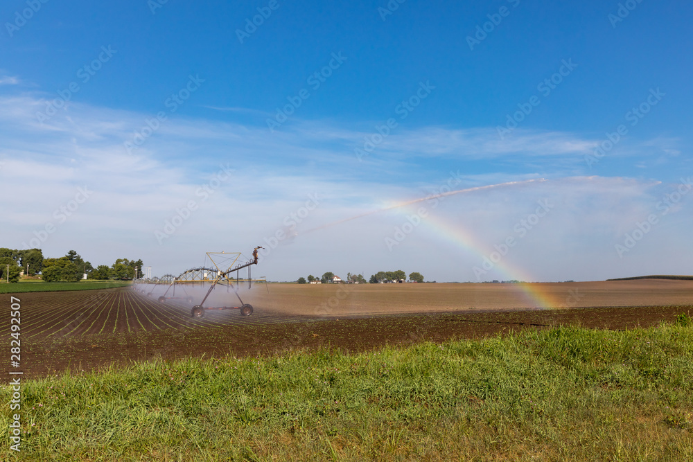 Center pivot irrigation system spraying water on recently planted soybean farm field creating a rainbow in the morning sun. Hot weather and little rain is creating drought conditions Illinois
