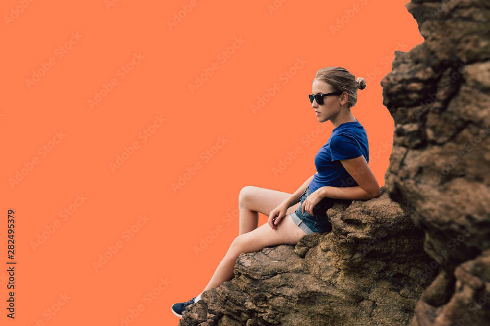 Young beautiful blonde tourist girl sits on a rocky ledge of a rock and looks far into the distance in the early foggy morning. Isolated human figure on the background of coral color.