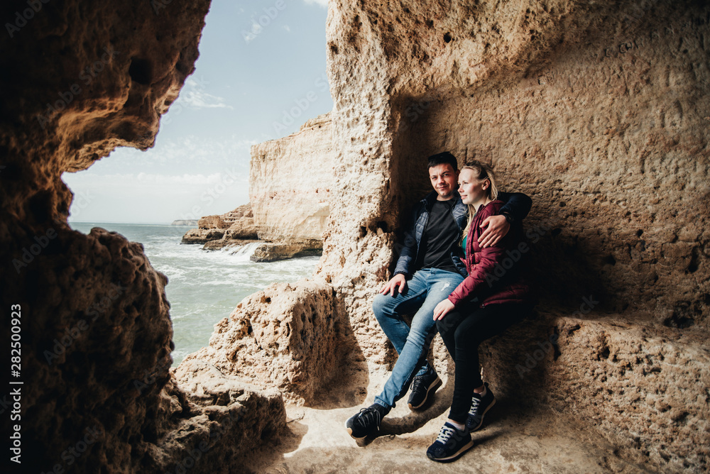 Happy young couple embracing and holding hands by the ocean view. Man and woman sitting together on the cliff. Smiling pair in black eyeglasses enjoying vacation in Portugal Happy young couple embracing and holding hands by the ocean view. Man and woman sitting together on the cliff. Smiling pair in black eyeglasses enjoying vacation in Portugal