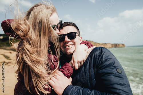 Happy young couple embracing and holding hands by the ocean view. Man and woman sitting together on the cliff. Smiling pair in black eyeglasses enjoying vacation in Portugal © Vadym
